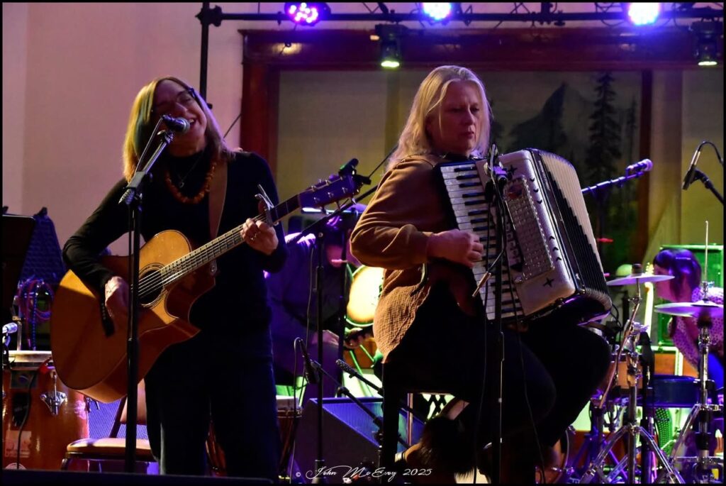 Annie Benjamin plays guitar, Jenni Mansfield Peal plays accordion on a stage.