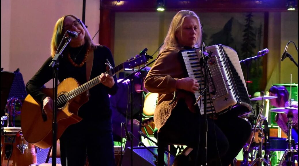 Annie Benjamin plays guitar, Jenni Mansfield Peal plays accordion on a stage.